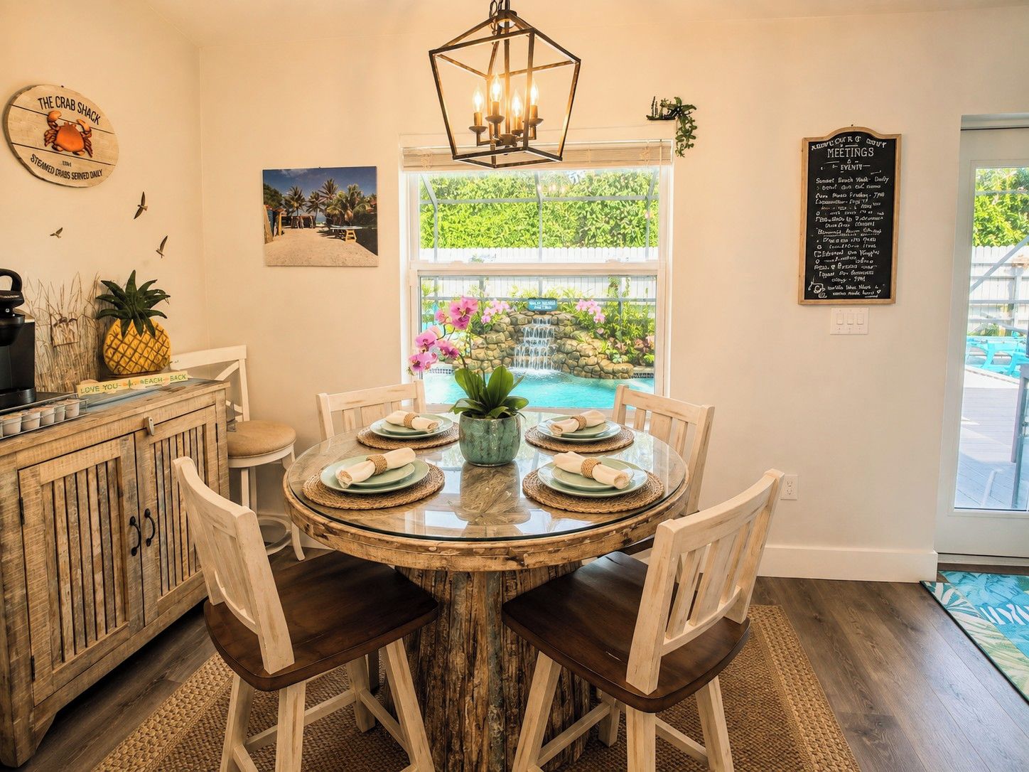 A dining area with a round table, chairs, a wooden cabinet, a hanging light fixture, a window with a view of greenery and a pool, and a chalkboard menu.