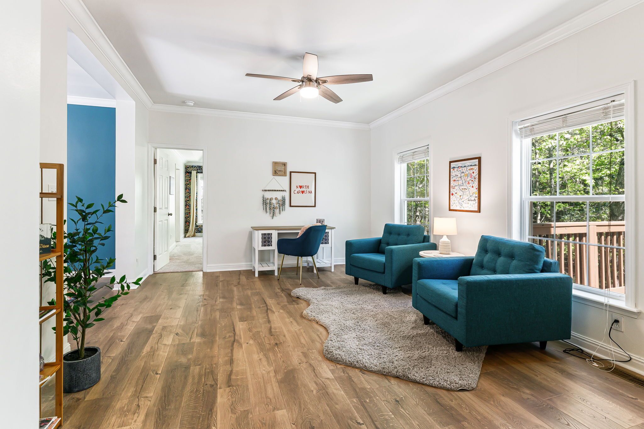 Un salon spacieux doté d’un plancher en bois, d’un ventilateur de plafond, d’un canapé bleu, d’un bureau avec une chaise et d’une grande fenêtre donnant sur les arbres.