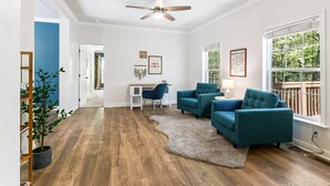 A spacious living room with a wooden floor, a ceiling fan, a blue sofa, a desk with a chair, and a large window with a view of trees. - 3 minutes to Pisgah Forest Entrance! - Game room - Fire pit   (Pisgah Forest)