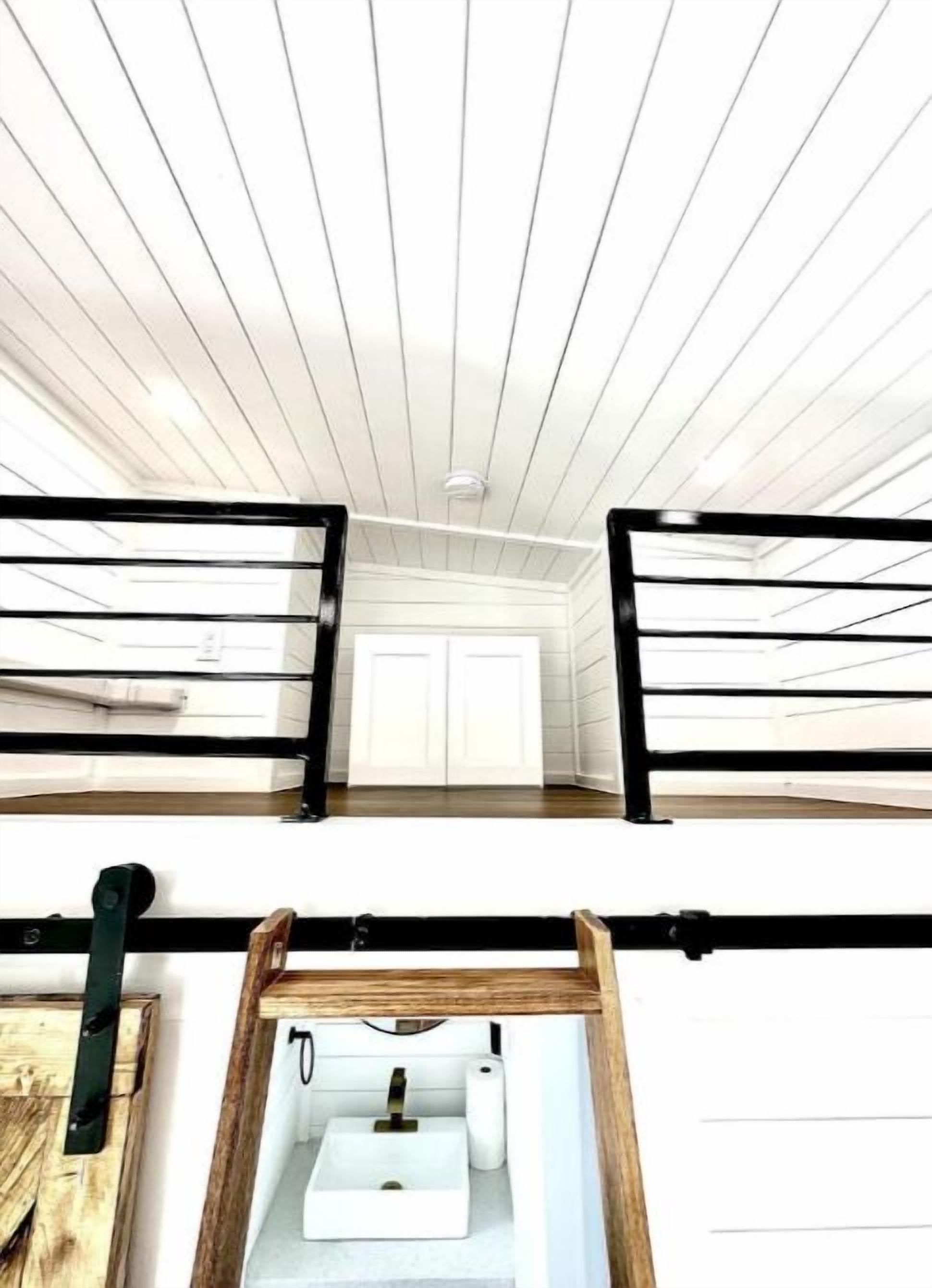 Ein Zimmer mit hohen Decken, einer weißen Decke und schwarzen Geländern. Ein weißes Waschbecken mit Wasserhahn und eine Toilettenpapierrolle sind sichtbar.