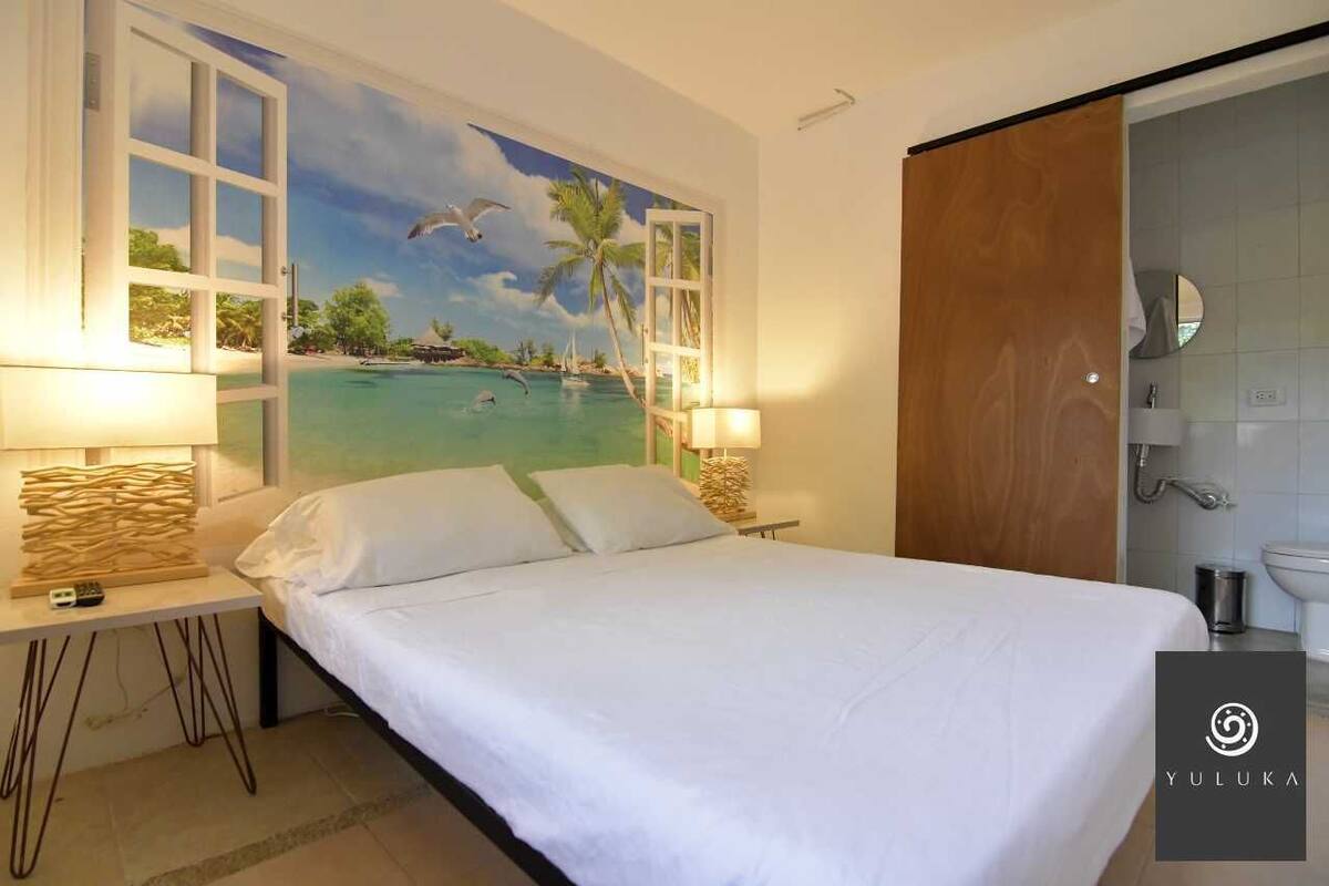 A hotel room with a bed, two bedside tables, a painting of a tropical beach, a wooden door, and a bathroom with a sink and mirror.