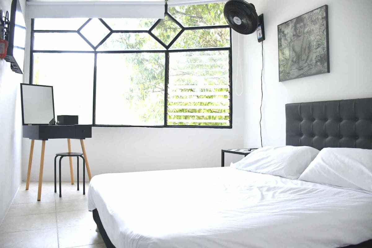 A bedroom with a bed, a desk with a chair, a window with a geometric design, and a painting on the wall.