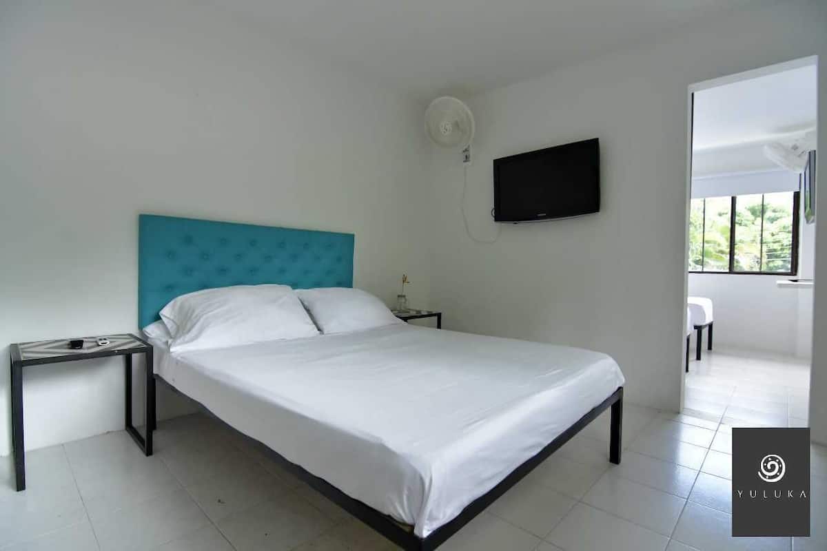 A modern hotel room with a single bed, a flat-screen TV mounted on the wall, and a small table with a lamp.