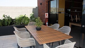Un área de comedor al aire libre con una mesa de madera y sillas de mimbre blanco, rodeada de plantas en macetas.