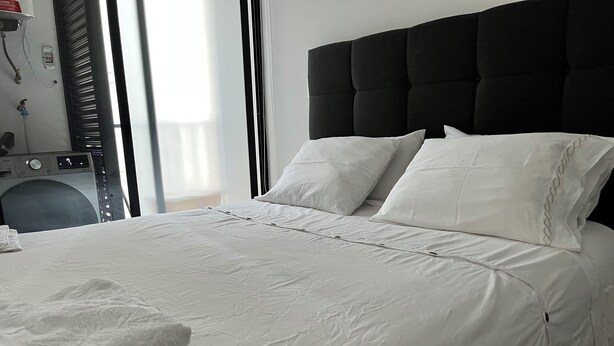 Una cama bien tendida con sábanas y almohadas blancas, cabecera negra y una pila de toallas blancas a la izquierda.