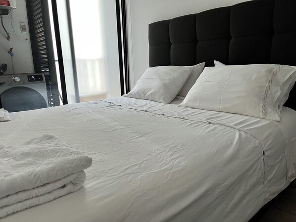 A neatly made bed with white linens and pillows, a black headboard, and a stack of white towels on the left side.