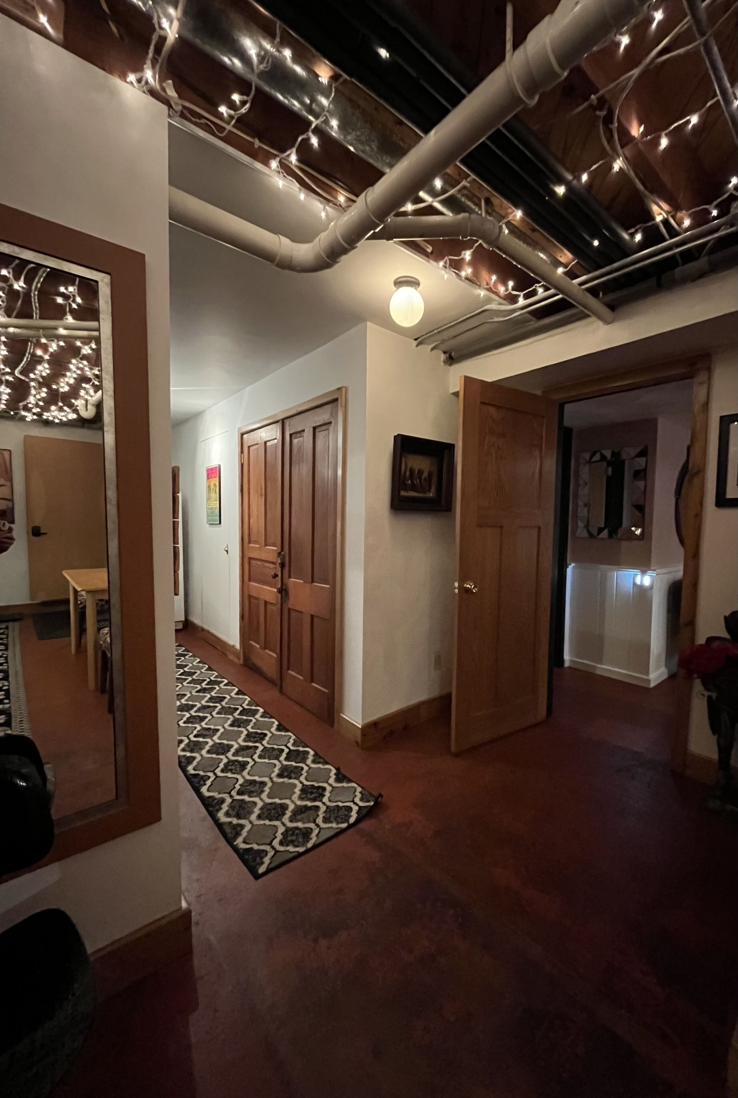 A hallway with wooden doors, a patterned rug, and exposed pipes with string lights.