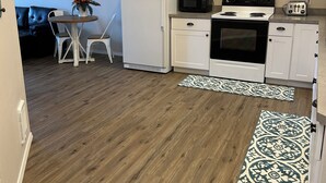 Cocina pequeña con armarios blancos, estufa negra, microondas y refrigerador. Hay una mesa de comedor con sillas y un adorno en la pared.