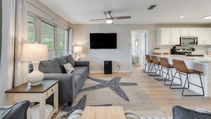 A modern living room with a gray sofa, a coffee table, and a TV. There is a kitchen area with a microwave, stove, and bar stools.