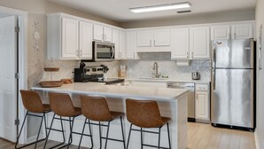 A modern kitchen with white cabinets, a stainless steel refrigerator, a double sink, and a bar area with stools.
