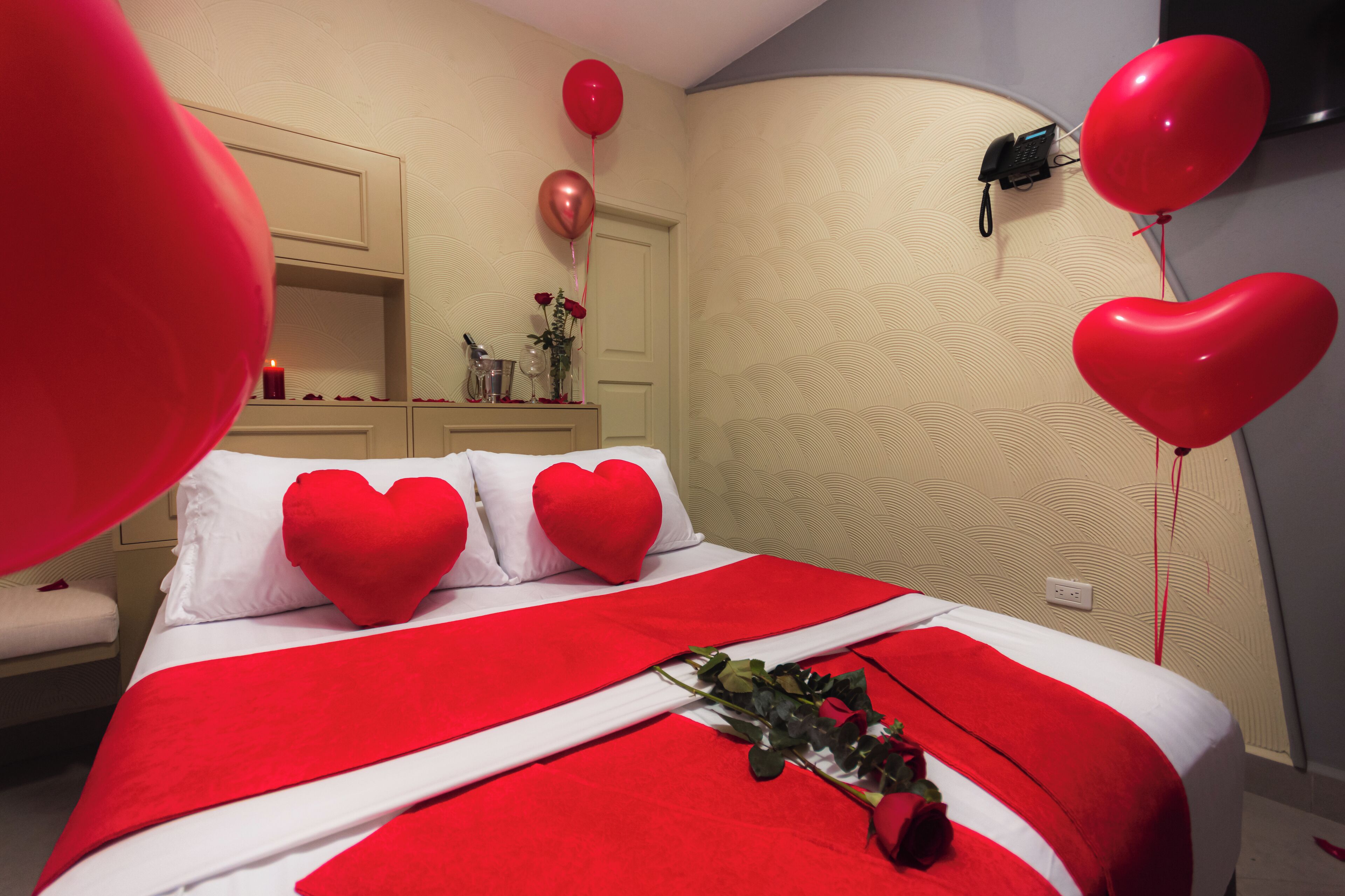 Quarto de hotel com cama, almofadas em formato de coração vermelho, um buquê de rosas vermelhas e balões vermelhos.