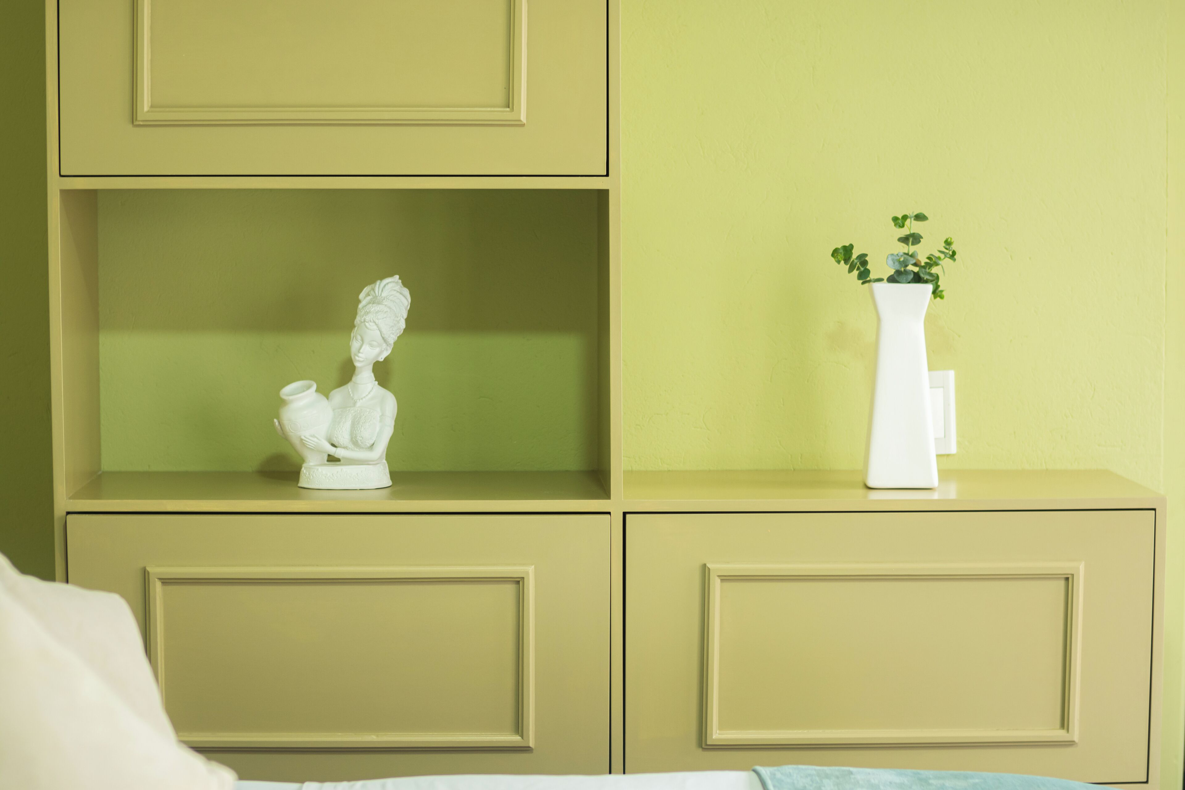 Una habitación con una cama, un jarrón blanco con una planta y un busto en una estantería.