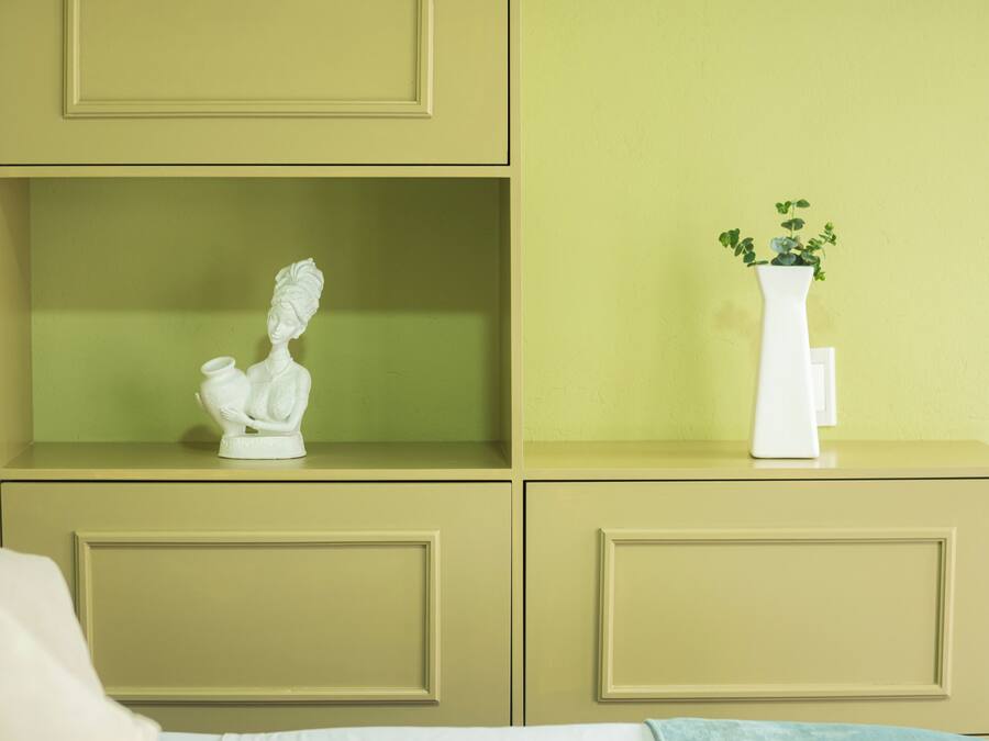 Una habitación con una cama, un jarrón blanco con una planta y un busto en una estantería.
