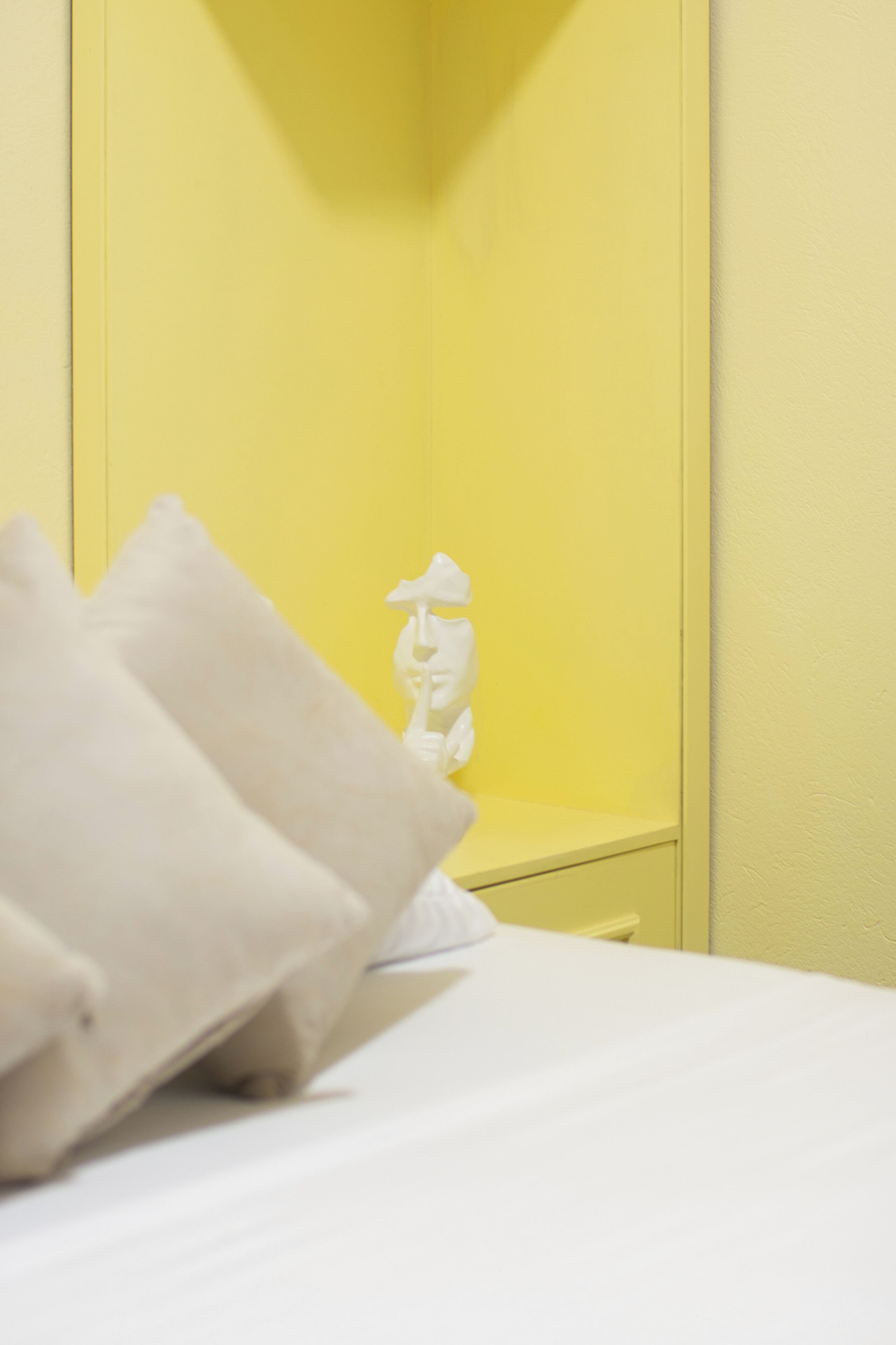 A bed with white bedding and pillows, a small white statue on a yellow nightstand, and a yellow wall.
