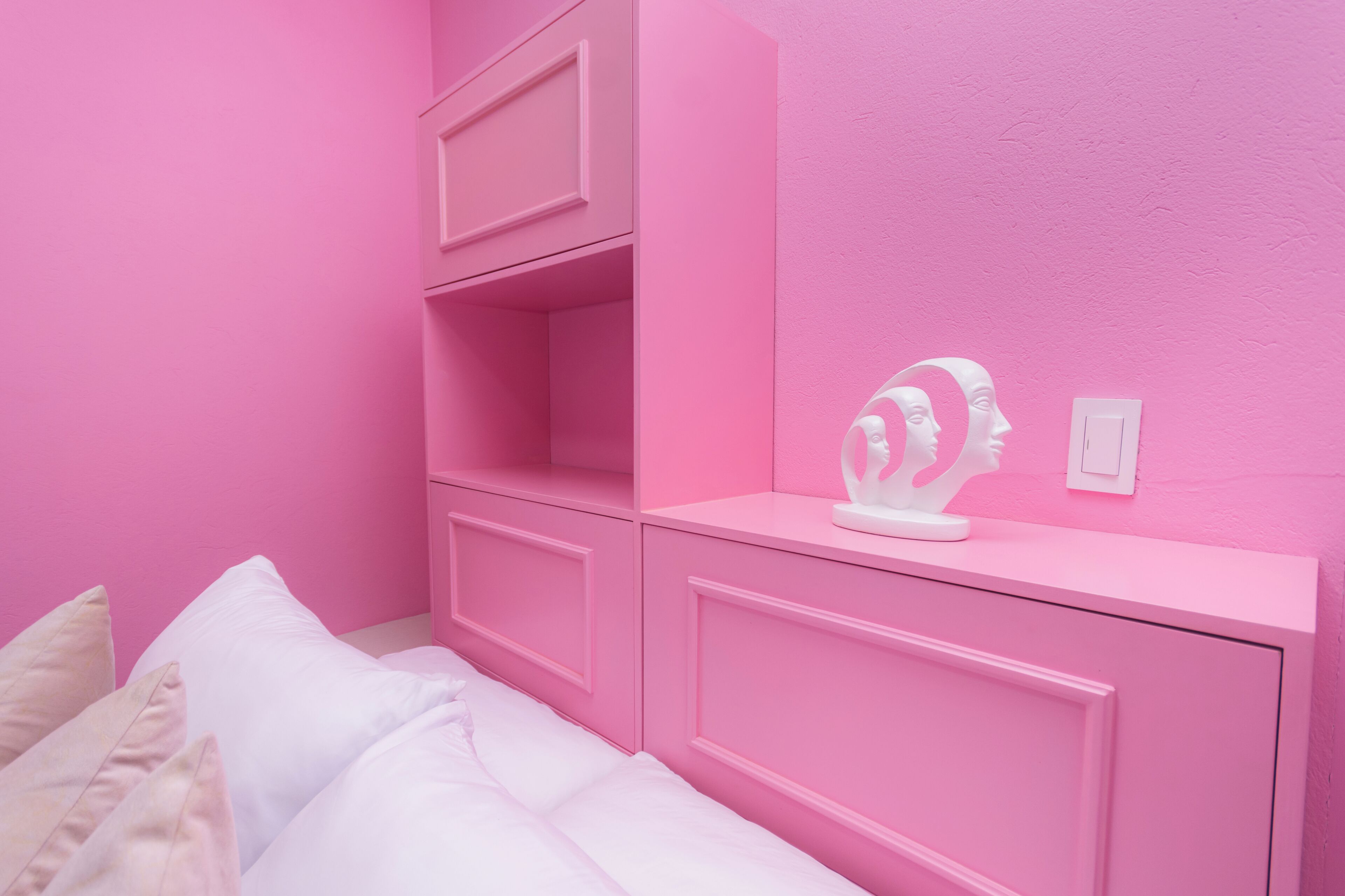 A pink-themed bedroom with a bed, white pillows, a dresser, and a wall-mounted sculpture.