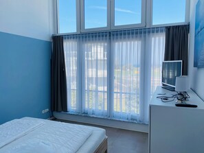 A room with a bed, a desk with a computer, a window with curtains, and a view of the outside.