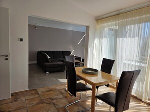 A modern living room with a sectional sofa, a dining table with chairs, and a large window with sheer curtains.