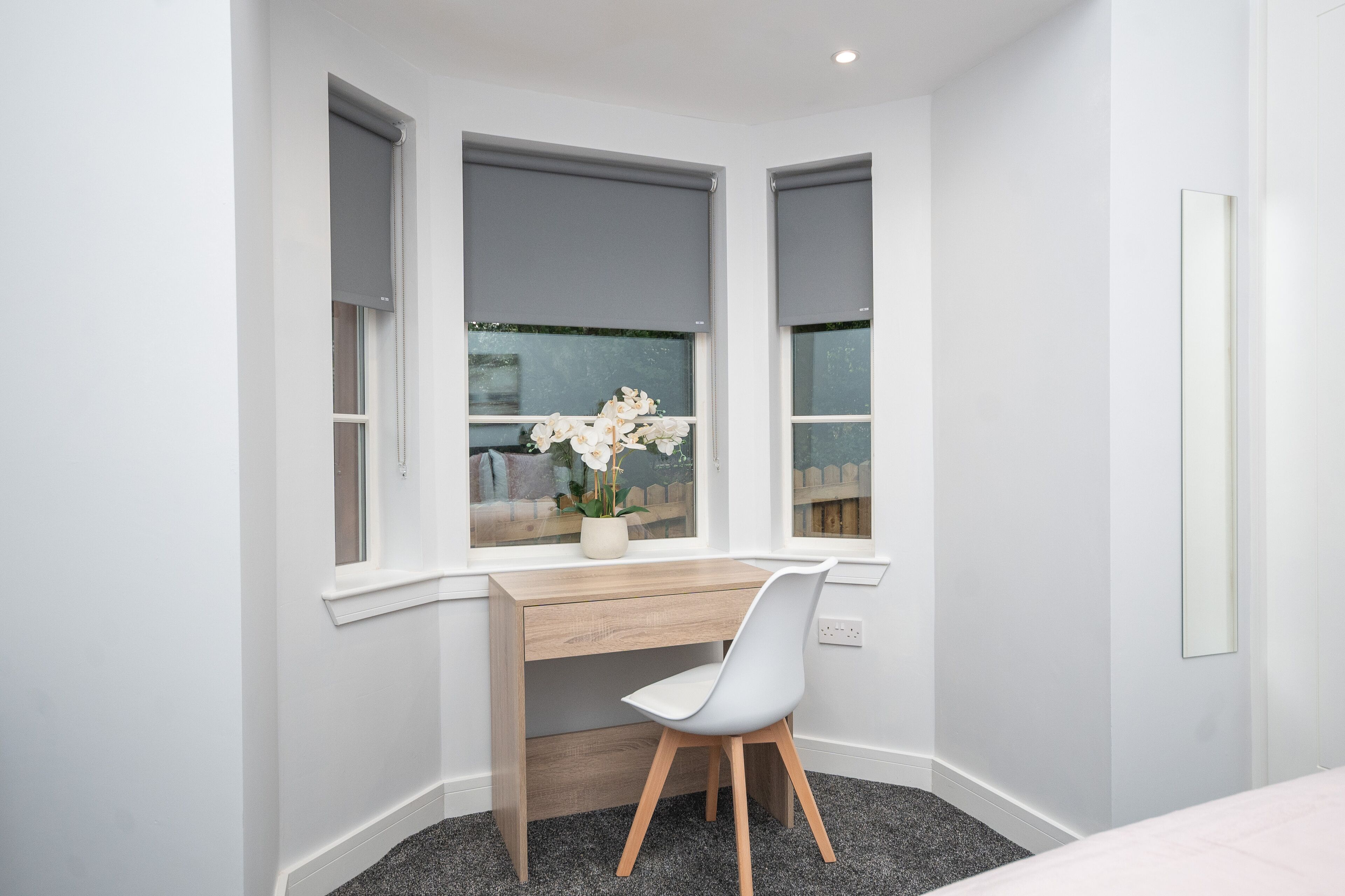 A corner desk with a white chair, a window with a view of a fence and flowers, and a mirror on the wall.