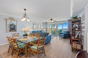 A spacious living room with a glass dining table, wicker chairs, a blue sofa, and a bookshelf. - Romar Tower 1A | Poolside | Gulf Front | My Beach Getaways (Orange Beach)