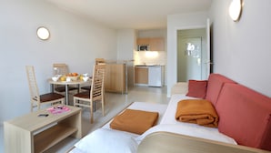 Une chambre d’hôtel de taille réduite comprenant un lit, un petit bureau avec une chaise, un coin repas avec une table et des chaises, et une kitchenette.