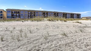 Un hotel de varios pisos con techo plano y grandes ventanales, ubicado en una playa de arena con poca vegetación.