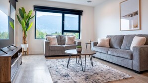 Un salon moderne avec un canapé gris, une table basse, un meuble pour la télévision et une grande fenêtre donnant sur une vue sur les montagnes.