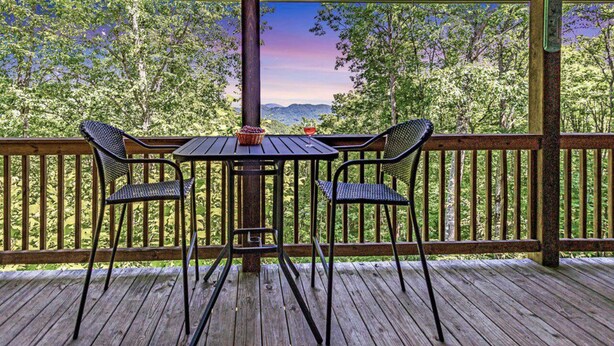 Una terraza de madera con una mesa y cuatro sillas, con vistas a una zona boscosa y montañas al fondo.