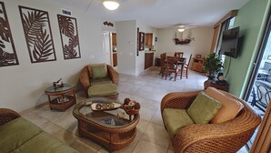 A spacious living room with wicker furniture, a dining area, and a kitchen. - 2br White Sands Village condo right at the beach (Kailua Kona)