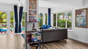 A modern living room with a grey sofa, a bookshelf, and a view of a pool and palm trees. - Fort Lauderdale~River Access~Kayaks~Heated Pool (Fort Lauderdale)
