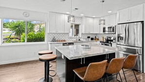 A modern kitchen with white cabinets, stainless steel appliances, a large island with bar stools, and a view of greenery outside. - Fort Lauderdale~River Access~Kayaks~Heated Pool (Fort Lauderdale)