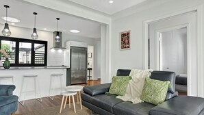 A modern living room with a black leather sofa, white bar stools, and a kitchen area in the background. - Modern family home in a leafy riverside suburb  (Graceville)
