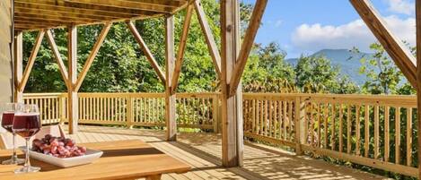 Une terrasse en bois avec une table et des chaises, une balustrade, et une vue sur la verdure et une montagne en arrière-plan.
