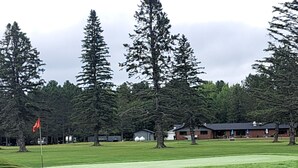 A golf course with a flag, tall trees, and buildings in the background. - 03 Dutchmen Classic that sleeps 1- 6 people comfortably for the perfect weekend  (Cloquet)