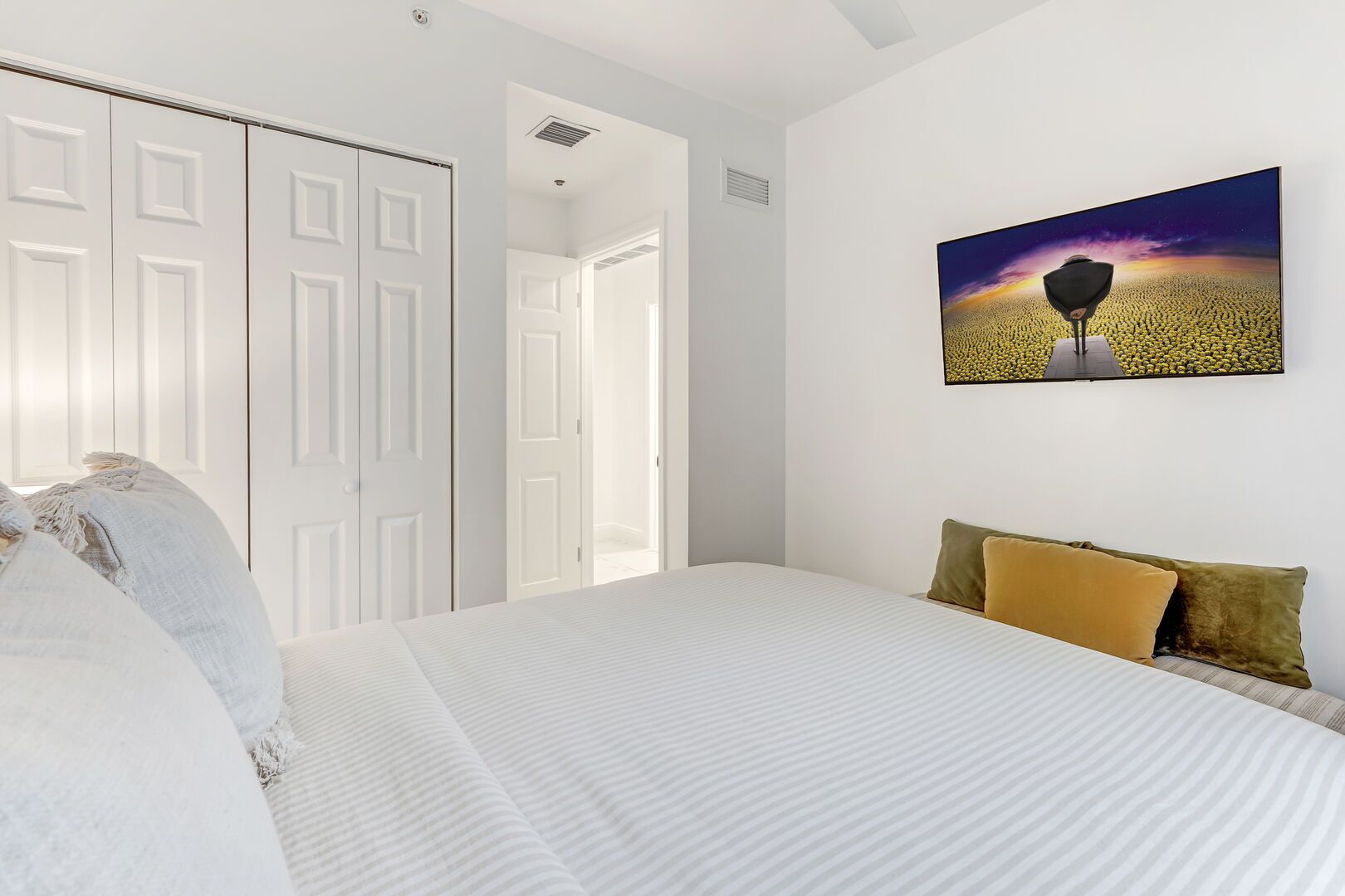 A bedroom with a large bed, white walls, a door, and a painting of a field.
