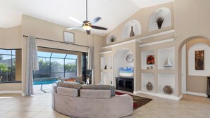 Ein geräumiges Wohnzimmer mit einer gebogenen Couch, einem Deckenventilator und einem großen Fenster mit Blick auf einen Swimmingpool.