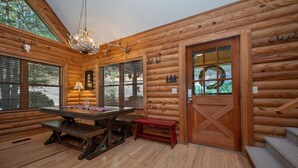 Interior de una cabaña de troncos con una mesa de comedor de madera, bancos, una araña y una puerta adornada con una corona.