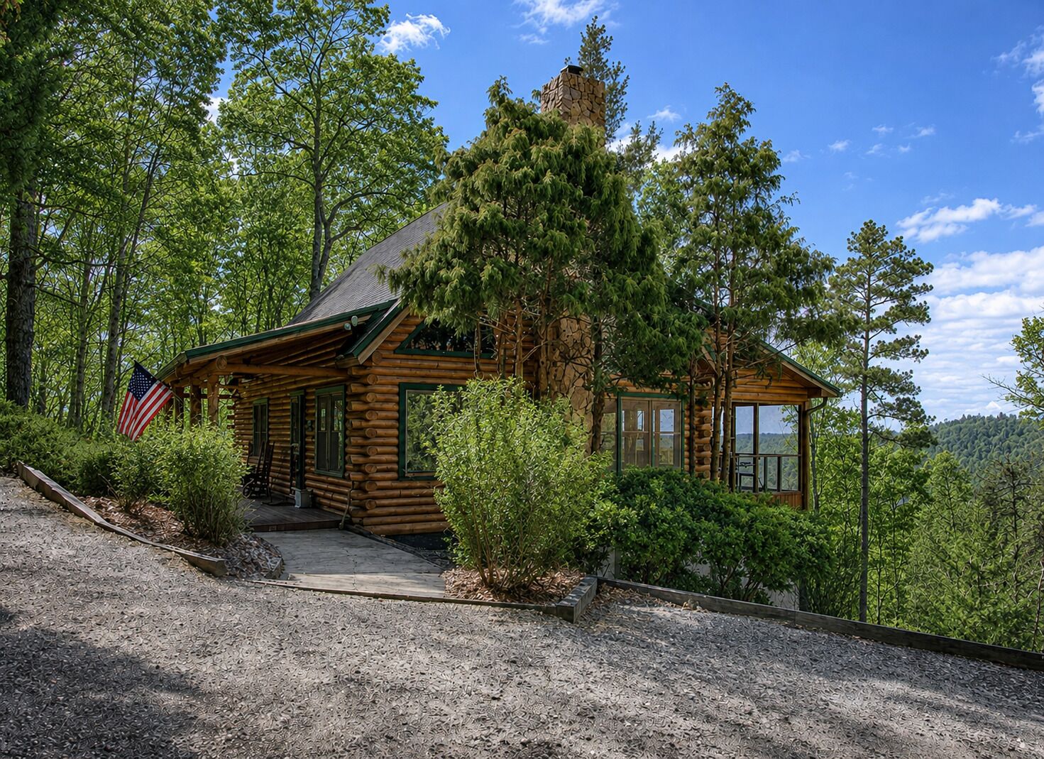 A log cabin with a stone chimney, surrounded by dense forest and a gravel driveway.
