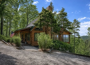 A log cabin with a stone chimney, surrounded by dense forest and a gravel driveway.