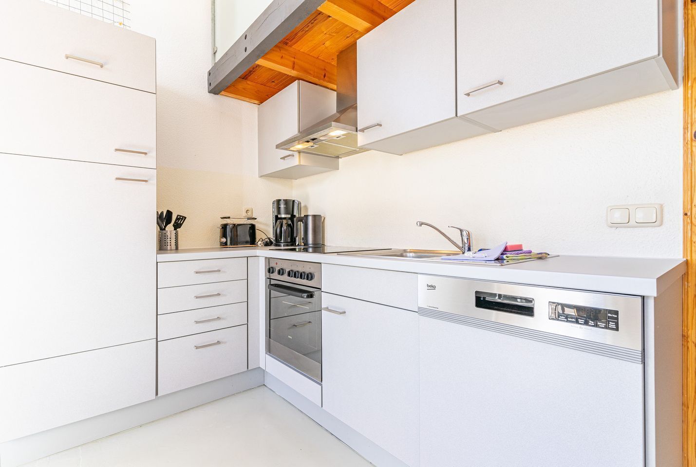 A modern kitchen with white cabinetry, a built-in oven, a dishwasher, and a coffee maker.