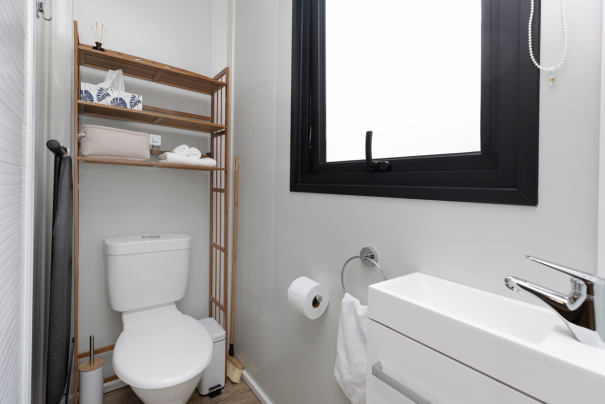 A compact bathroom with a toilet, a shelf with towels, a window, and a sink.