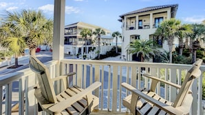 A balcony with two wooden chairs, a white railing, and a view of a multi-story building with balconies and palm trees.