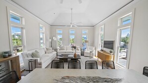 A spacious living room with a large central table, white sofas, and a TV mounted on the wall.