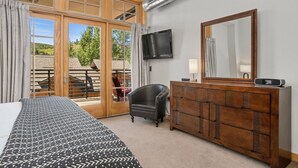 Une chambre d’hôtel avec un lit, une commode en bois, une chaise, un téléviseur et une grande fenêtre donnant sur une terrasse en bois et des arbres.