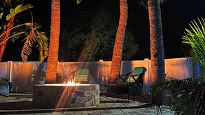 An outdoor patio area with a fire pit, two chairs, and tall palm trees. - Pool Paradise with hot tub, bikes, every beach need, games and more! (Cocoa Beach)