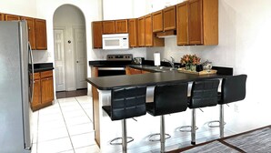 A modern kitchen with wooden cabinets, a stainless steel refrigerator, a microwave, an oven, and a bar area with four black bar stools. - Pool Paradise with hot tub, bikes, every beach need, games and more! (Cocoa Beach)
