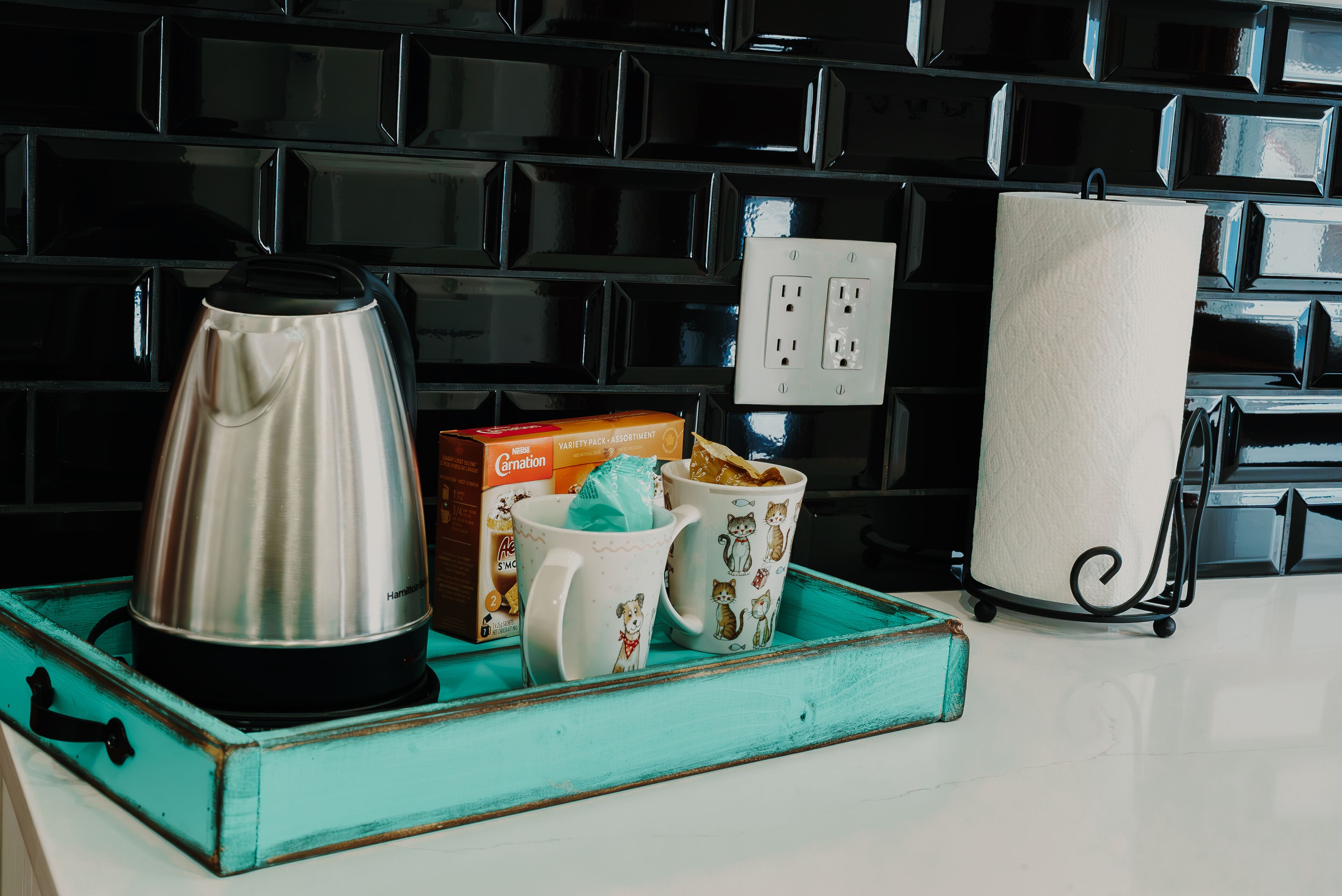 A kitchen counter with a stainless steel kettle, a box of tea, two mugs with cat designs, and a paper towel holder.