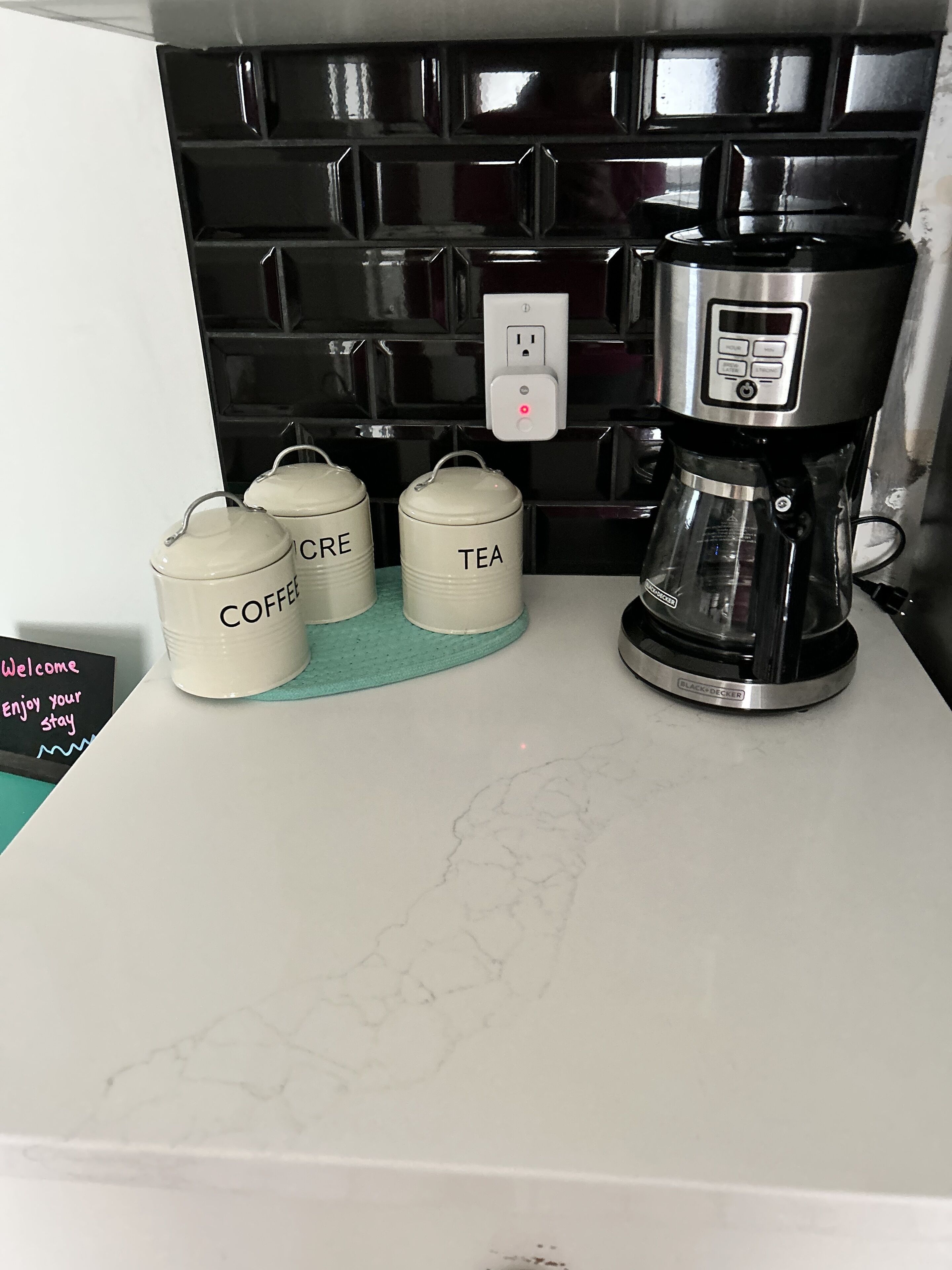 A kitchen counter with a coffee maker, two containers labeled 'COFFEE' and 'TEA', and a welcome sign.
