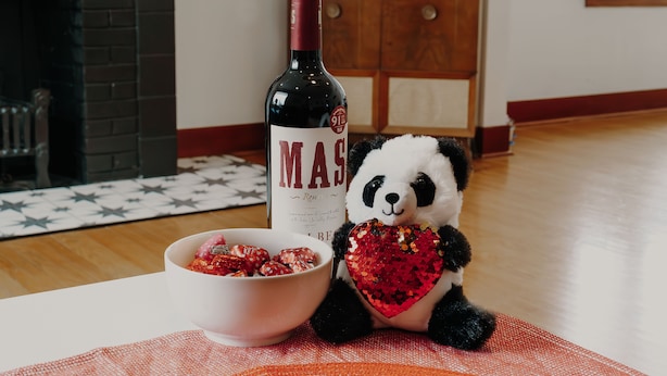 Ein kuscheliges Panda-Spielzeug mit herzförmigem Zubehör, eine Weinflasche und eine Schüssel mit Snacks auf einem Tisch.