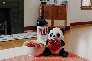 Un peluche de panda con un accesorio con forma de corazón, una botella de vino y un bol de aperitivos sobre una mesa.