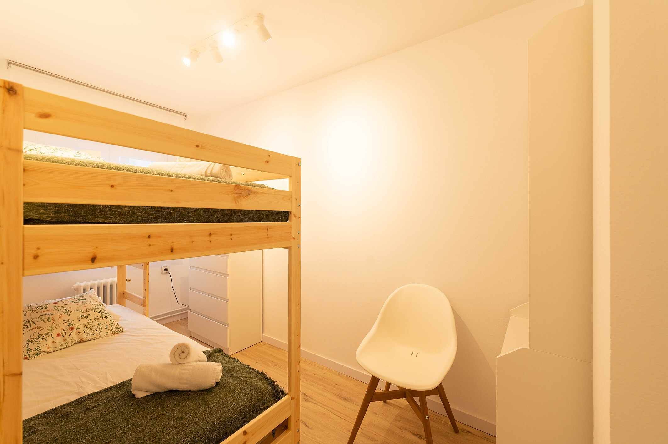 A bunk bed with a green mat, a white chair, and a white dresser in a room with white walls and wooden flooring.